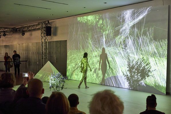 Ein Publikum sitz in einem verdunkelten Raum vor einer Leinwand, die mit abstrakten Formen und buntem Licht beleuchtet wird. Eine Person inszeniert ein Performance. 