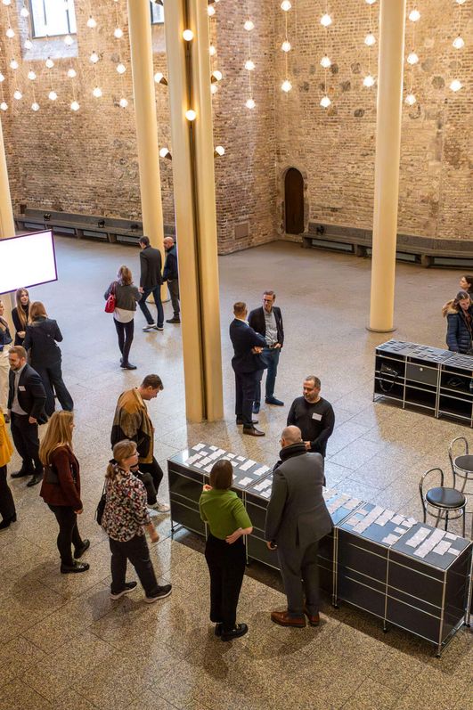 Das Foyer des Gürzenichs aus der Vogelperspektive. Menschen stehen verteilt im großzügigem Raum. Von der Decke hängen einzelne Glühbirnen, die dem Raum eine warme Atmosphäre verleihen.