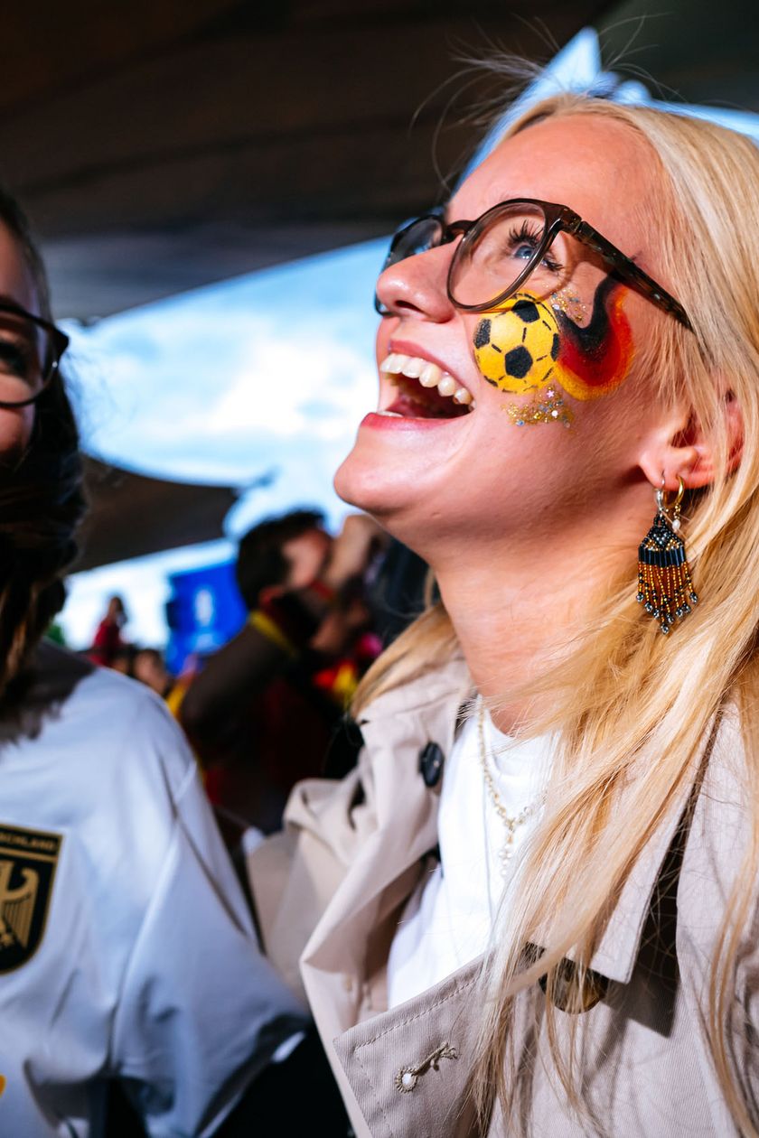 Zwei Frauen in Trikots sind fröhlich und lachen. Flaggen verzieren ihre Wangen. 
