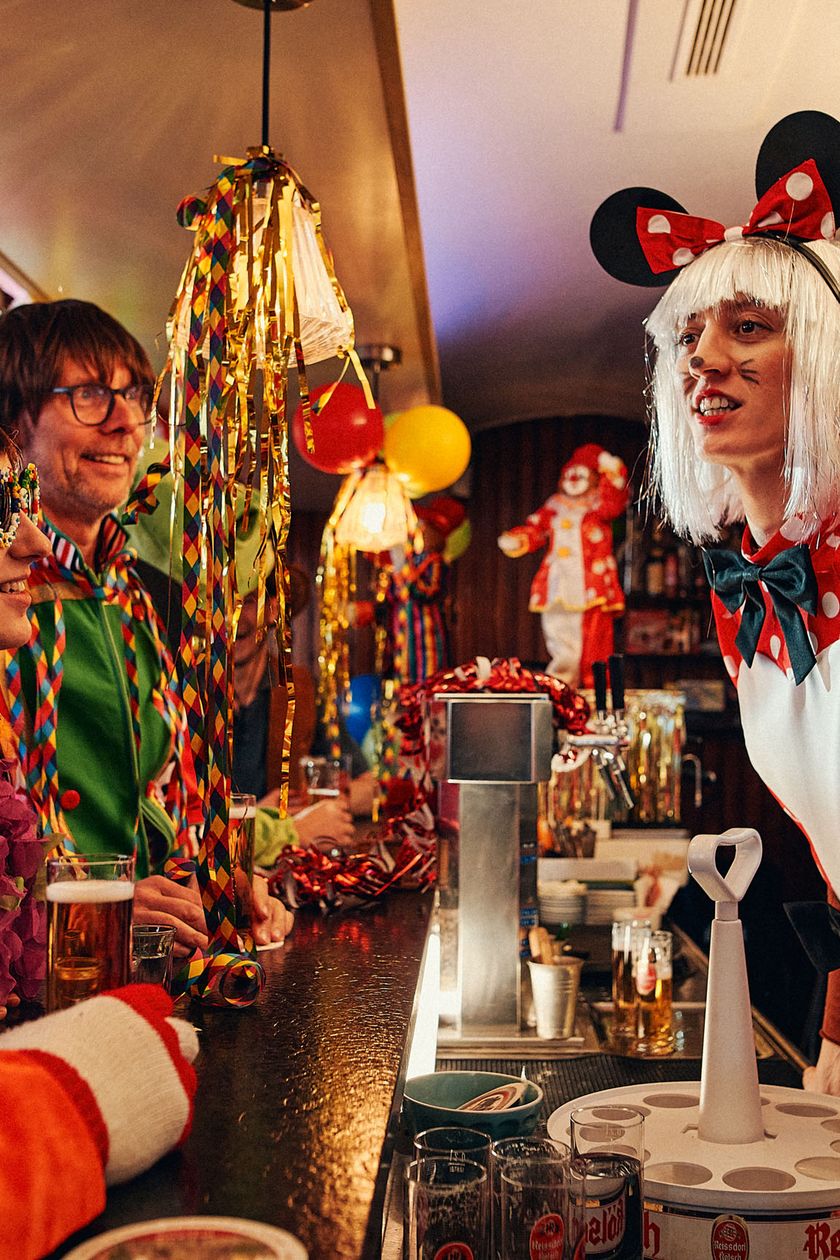 Zwei verkleidete Frauen stehen nach vorne gelehnt hinter einer Bartheke. Sie sind im Gespräch mit drei Personen, die vor der Bar stehen. Luftballons, Luftschlangen und Lametta hängen an der Decke und hüllen den Raum eine bunte und fröhliche Atmosphäre.