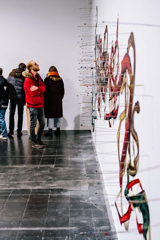 Auf einer weißen Wand ist ein geschwungenes Wandgemälde gemalt wodrauf viele dünne Stäbe fixiert sind. Personen schauen sich das Kunstwerk an.