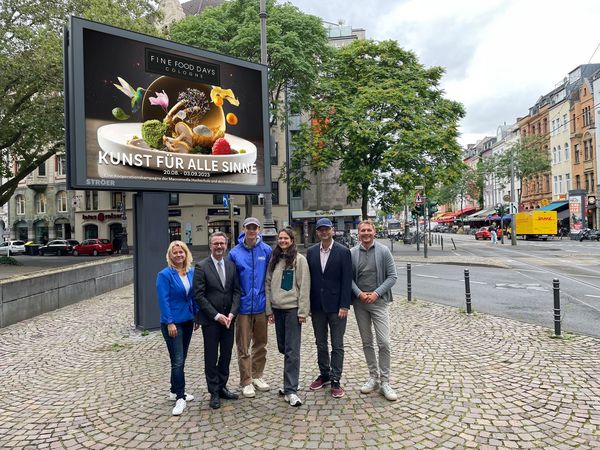 Fünf Personen posieren auf einem kleinen Platz mit Pflastereinen vor einem großen Ströer Bildschirm. Im Hintergrund führt einen Straße gesäumt von Altbauten.