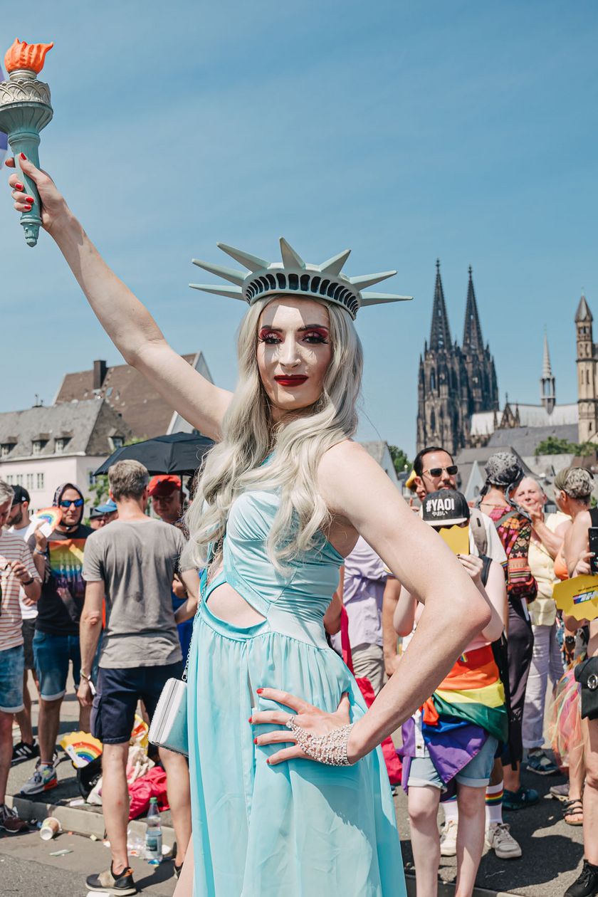 Eine Person verkleidet als Freiheitsstatue posiert. Im Hintergrund stehen Menschen am Wegrand.