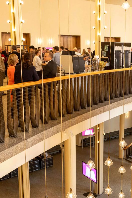 Auf dem Bild ist das Foyer und obere Etage vom Gürzenich zu sehen. Menschen unterhalten sich oder bedienen sich am Buffet.