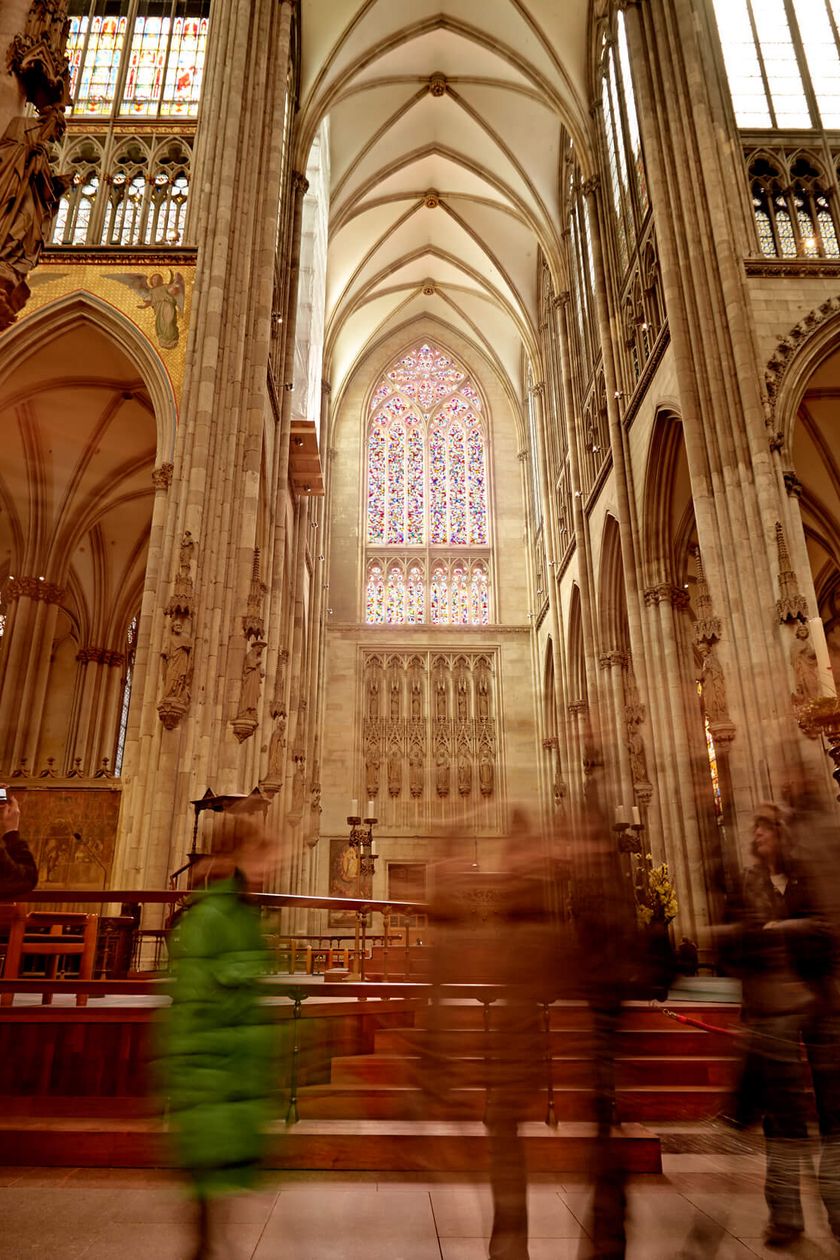 Licht bricht durch das Richterfenster im Kölner Dom. Menschen bewegen sich durch den Innenraum vom Kölner Dom.