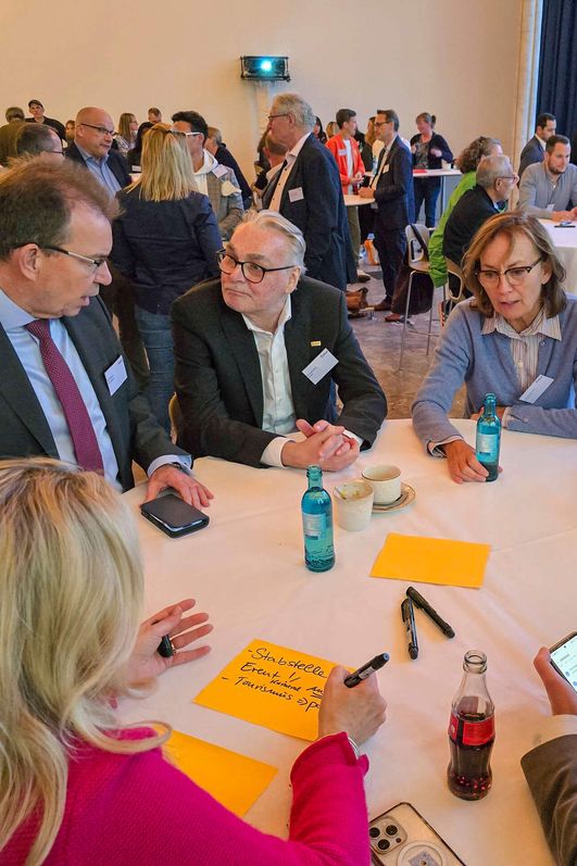 Um einen runden Stehtisch erarbeitet eine Gruppe von Menschen eine Aufgabe und schreibt ihre Gedanken auf Notizzettel. 