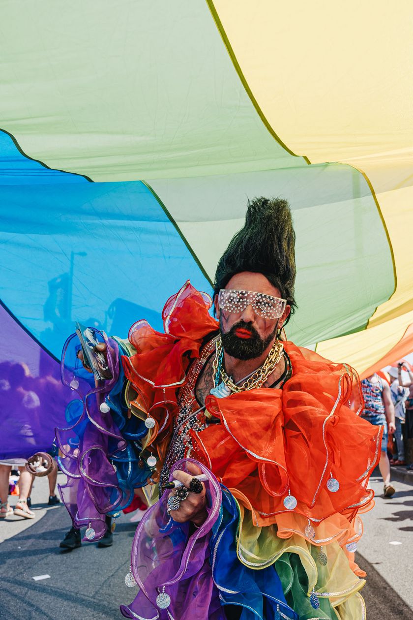 Eine Person in farbenfrohem Kostüm posiert. Über ihr eine Regenbogenfahne ausgebreitet.