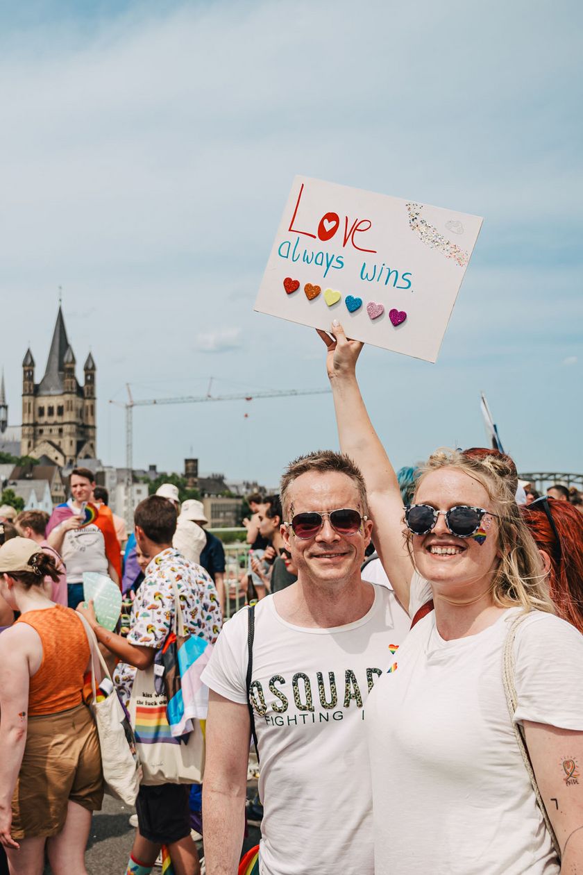 Zwei Personen posieren und halten eine Schild in die Höhe. Menschen laufen dran vorbei und im Hintergrund ist der Kölner Dom zusehen.