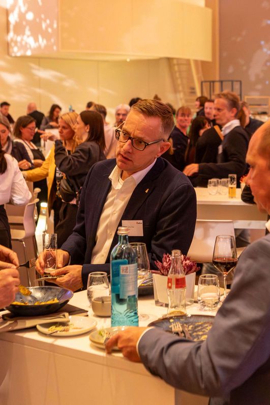 Vier Männer stehen um einen Stehtisch und verzehren Speisen. Dabei unterhalten sie sich untereinander.