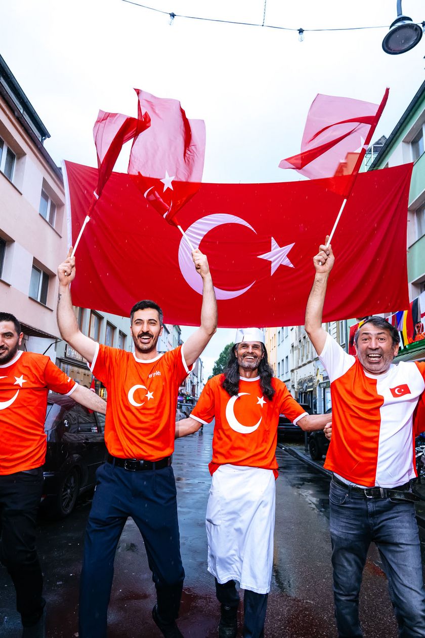 Sechs Männer in Trikots laufen eine Straße hinunter und halten eine große Flagge in die Luft. 