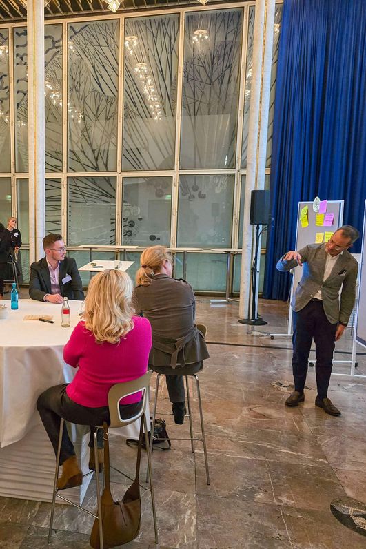 Ein Moderator erklärt vor einem Publikum eine Thematik anhand von Notizen, die an Flipcharts befestigt sind.