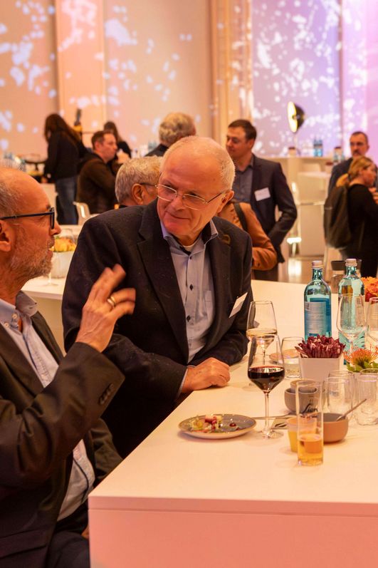 Ein belebter Saal, gehüllt in warmes Licht. Menschen unterhalten sich oder essen an Stehtischen. 