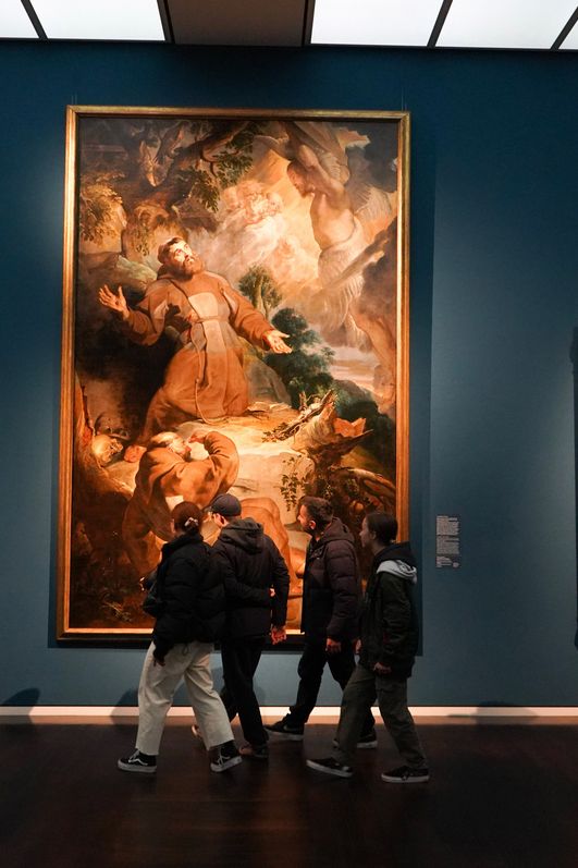 Vier Personen gehen an einem großen Gemälde mit goldenem Rahmen vorbei. Weitere Gemälde hängen an den dunkelblau gestrichenen Wänden.