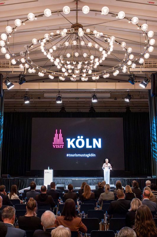 Eine Sprecherin steht vor einem Publikum auf einer Bühne und hält einen Vortrag.