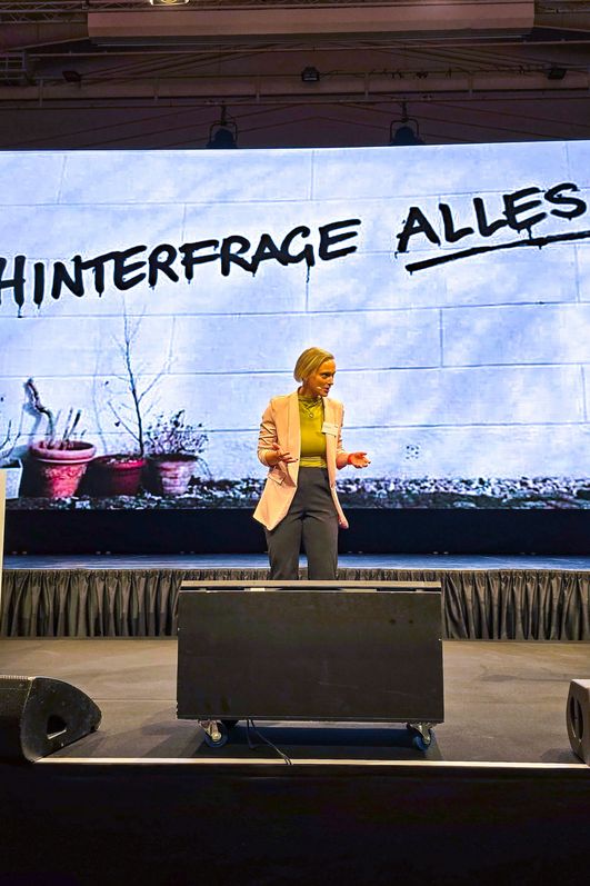 Eine Moderatorin steht auf der Bühne und spricht zu einem Publikum. 