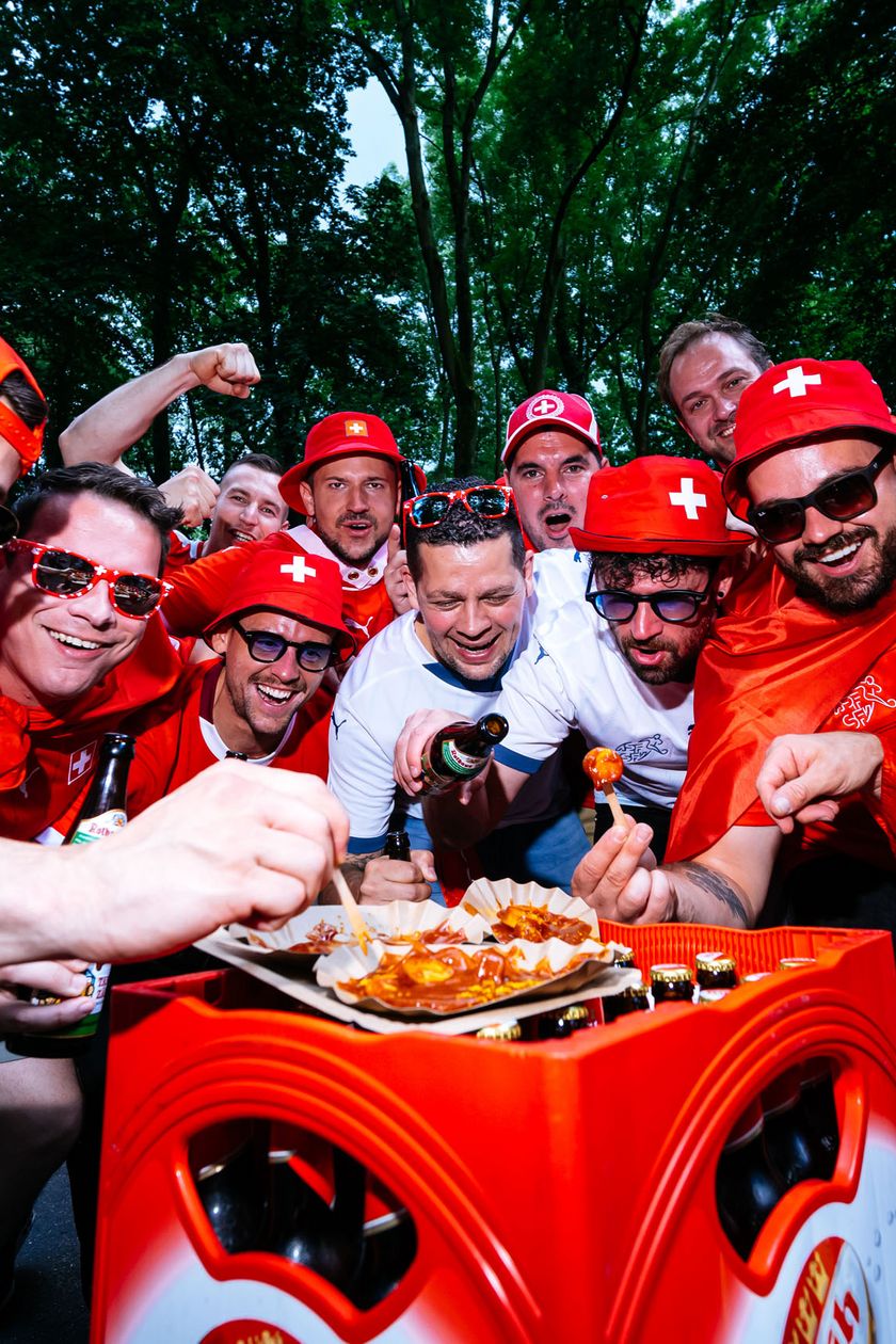 Eine Gruppe von Männern in roten Trikots steht um einen Kastenbier herum. Darauf eine Pappschüssel Currywurst mit Pomms. Die Männer stechen mit einer Gabel in das Gericht und sehen dabei fröhlich aus.