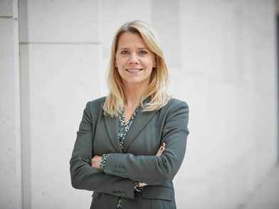 Frau Stephanie Kleine Klausing posiert selbstsicher mit freundlichem Lächeln vor einer weißen Mauerwand.