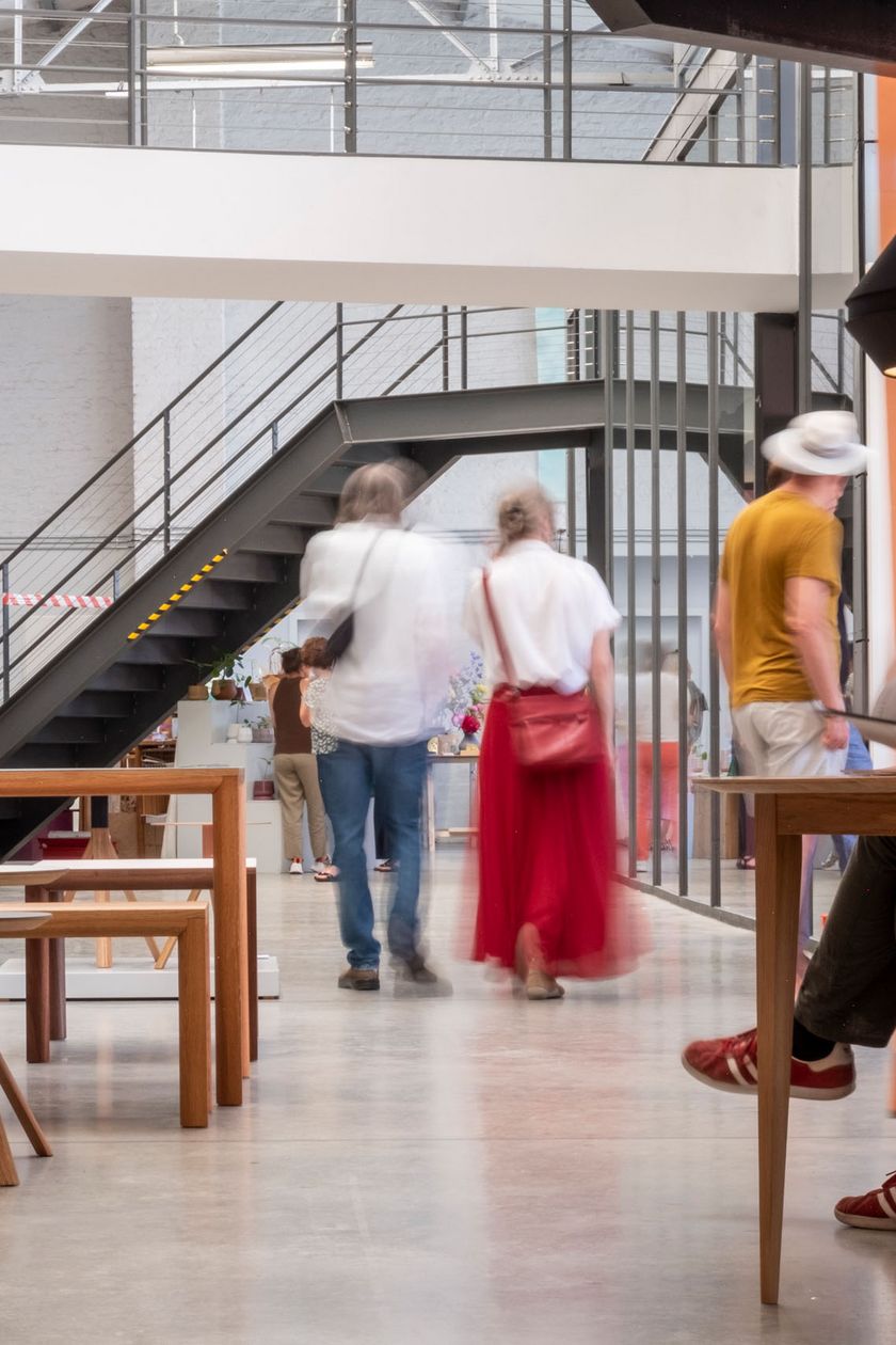 Auf dem Bild ist eine hell mit lichtdurchflutete Halle zusehen, mit einem Brückensteck und Treppenaufgang. Designmöbel sind in der Halle platziert und Menschen schauen sich interessiert um.