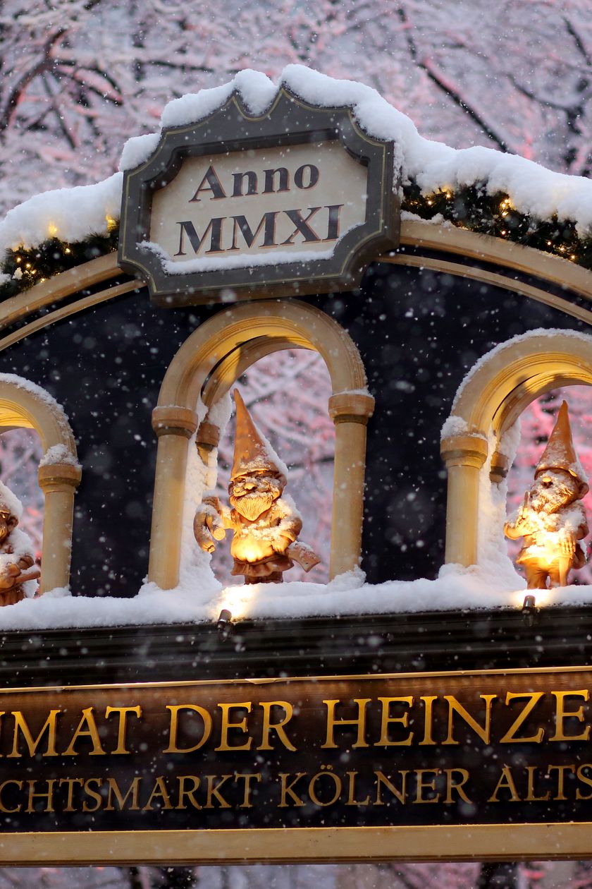 Der Eingang zum Kölner Weihnachtsmarkt am Heumarkt bei Abenddämmerung. Auf dem Bild ist der verzierte Torbogen mit drei Heinzelmännchenfiguren zu sehen, gehüllt mit Schnee.