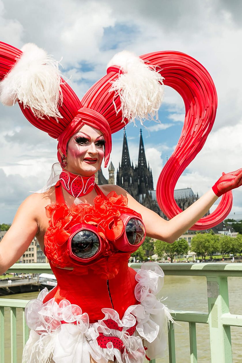 Eine Person in einem phantasievollen rotem Kostüm posiert auf der Deutzer Brücke, im Hintergrund erhebt sich der Kölner Dom.