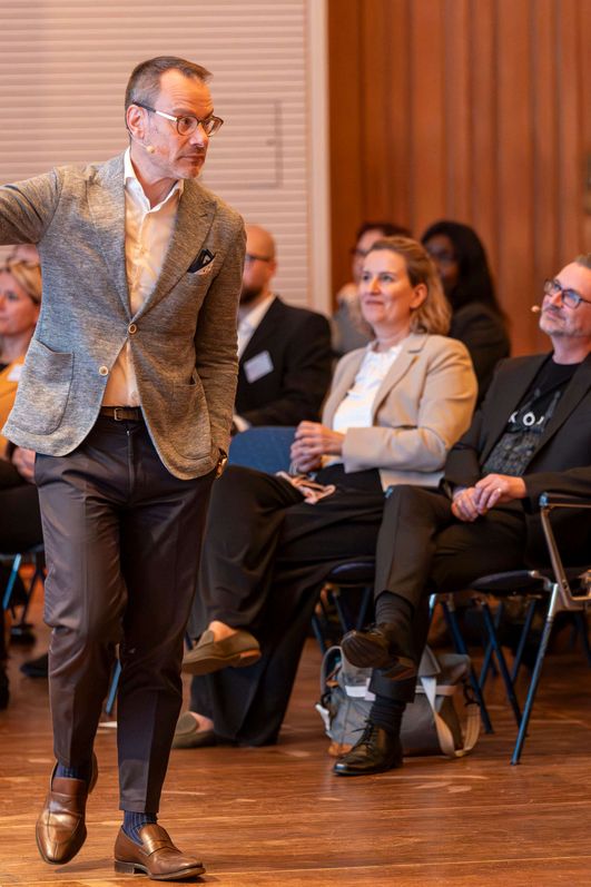 Ein Sprecher steht direkt vor einem Publikum und zeigt in eine Richtung. 