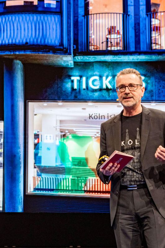 Herr Dr. Amann steht vor einer Bildschirmpräsentation, die ein Bild der Außenansicht der Tourist Info abbildet.