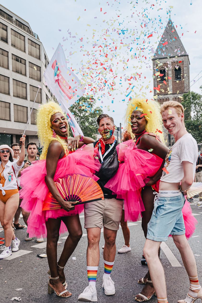 Vier Personen posieren bei der Pride Demonstration auf der Straße. Zwei Personen tragen blonde Perücken und pinke Tüllröcke. 