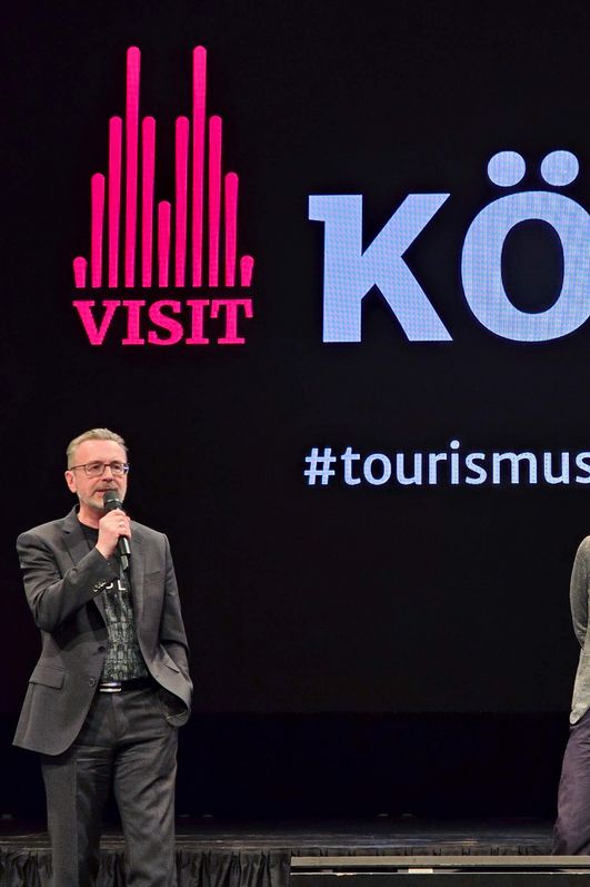 Herr Dr. Amann spricht in ein Mikrofon auf einer Bühne. Zwei weitere Personen stehen daneben und hören aufmerksam zu.