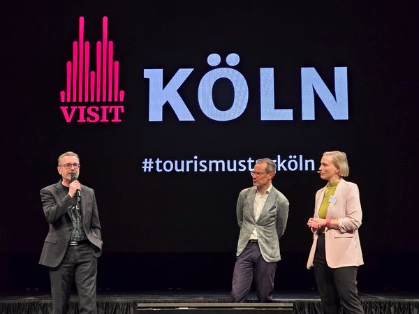 Herr Dr. Amann spricht in ein Mikrofon auf einer Bühne. Zwei weitere Personen stehen daneben und hören aufmerksam zu.