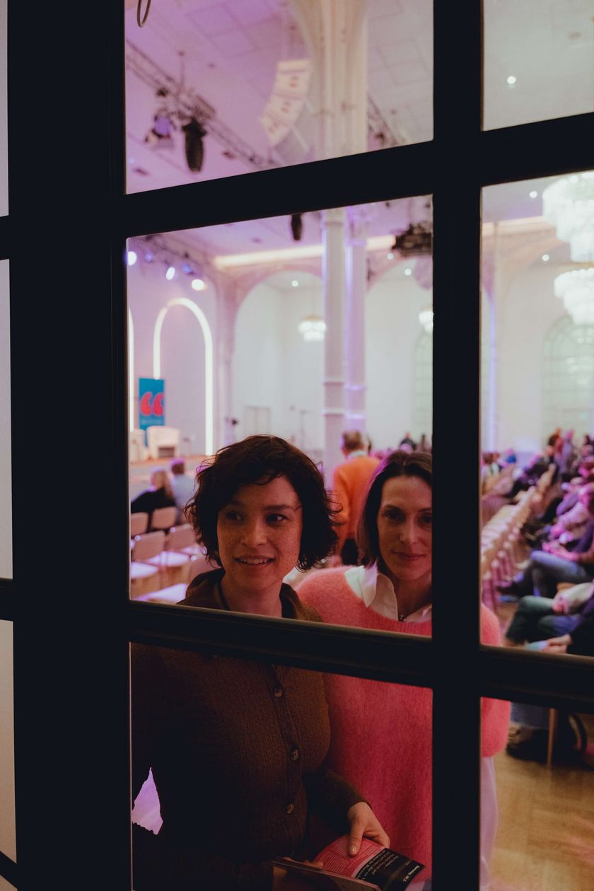 Zwei Personen schauen durch eine Glastür. Hinter ihnen ein warm beleuchteter Saal.