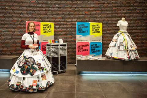 Vor einer dunklen Kachelwand steht eine Frau mit kreativem ausgestelltem Rock aus Zeitungspapier. Neben ihr sind Aufsteller mit Reklame zu sehen und ein Schaufensterpuppe auch mit einem Zeitungspapierrock. 
