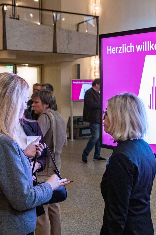 Auf dem Bild ist das Foyer des Gürzenich zu sehen. Besucher*innen unterhalten sich. Ein Bildschirm steht im Vordergrund vom Bild, darauf zu lesen "Herzlich Willkommen. Kölner Tourismustag".
