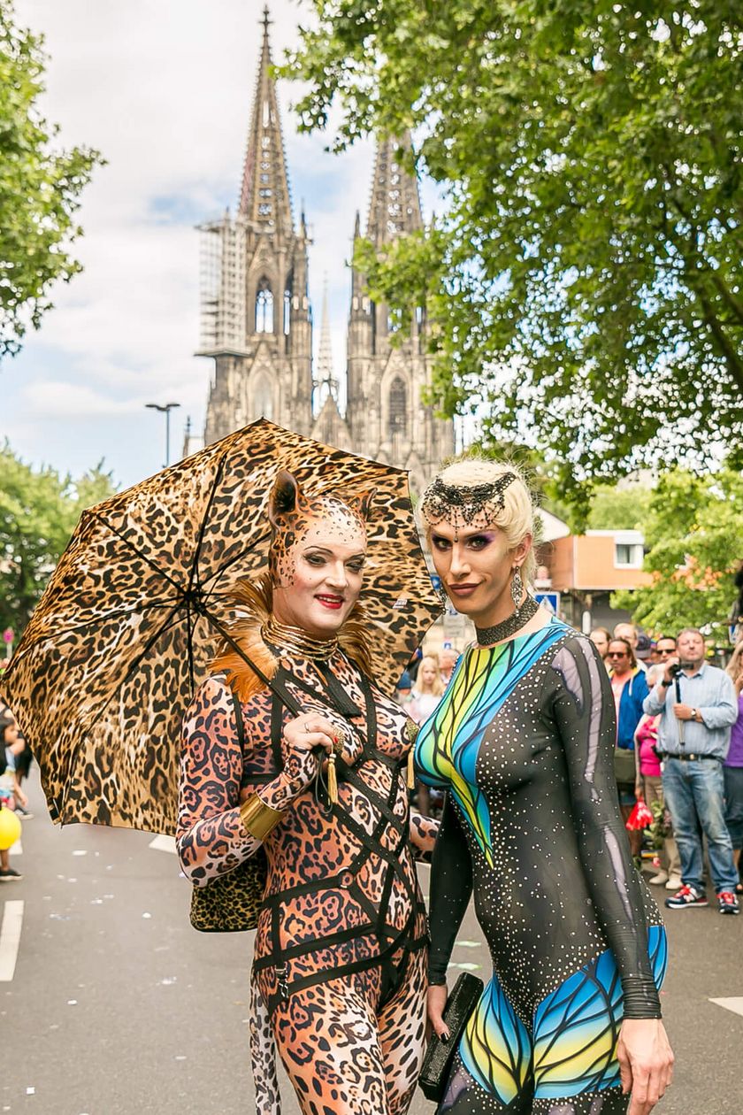 Zwei Personen in kreativvollen Kostümen posieren auf dem Weg der Pride Demonstration. Menschen stehen am Wegrand und im Hintergrund erstreckt sich der Kölner Dom.