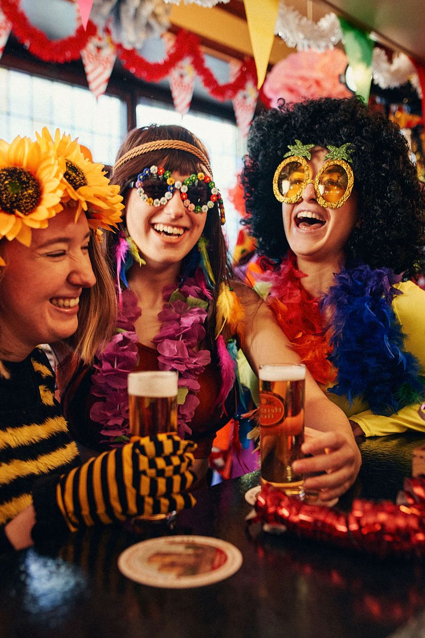 Drei verkleidete Frauen mit Sonnenblumen in den Haaren, großen runden Brillen, stehen in einem farbenfroh geschmückten Raum an einer Bartheke und lachen ausgelassen.