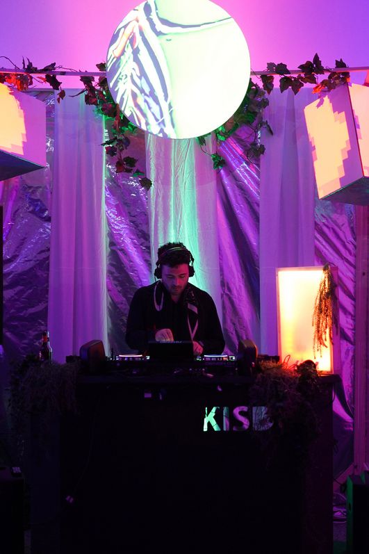 In einem lila beleuchteten Raum steht mittig ein DJ Pult und eine Person steht davor. Der Hintergrund ist mit glänzenden und glitzernden Materialien dekoriert.