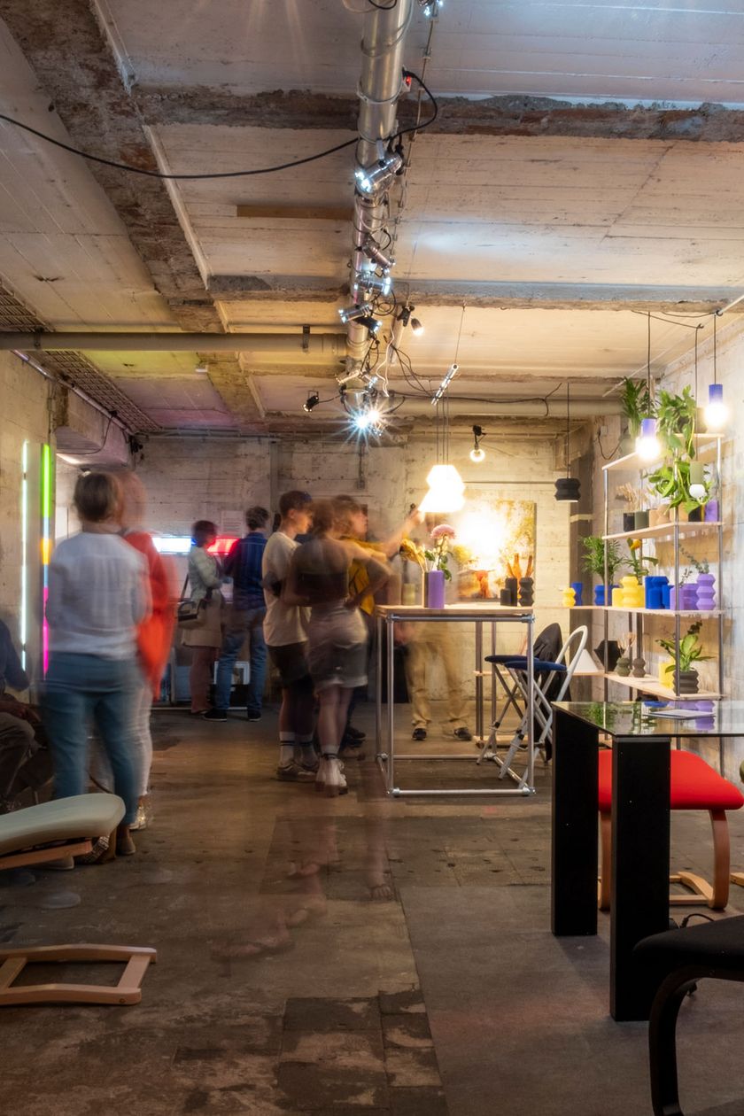 In einem unterirdischen industriellen Raum mit Betonwänden sind Designmöbelstücke und Dekoartikel verteilt. Menschen schauen sich die Ausstellung an.