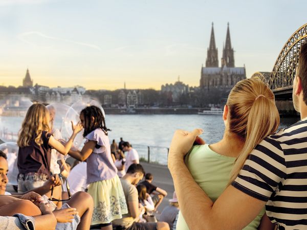 Eine Gruppe von Menschen genießen den Sonnenuntergang am Rheinboulevard mit Blick auf den Kölner Dom und die Altstadt. Die Menschen interagieren nicht miteinander. 