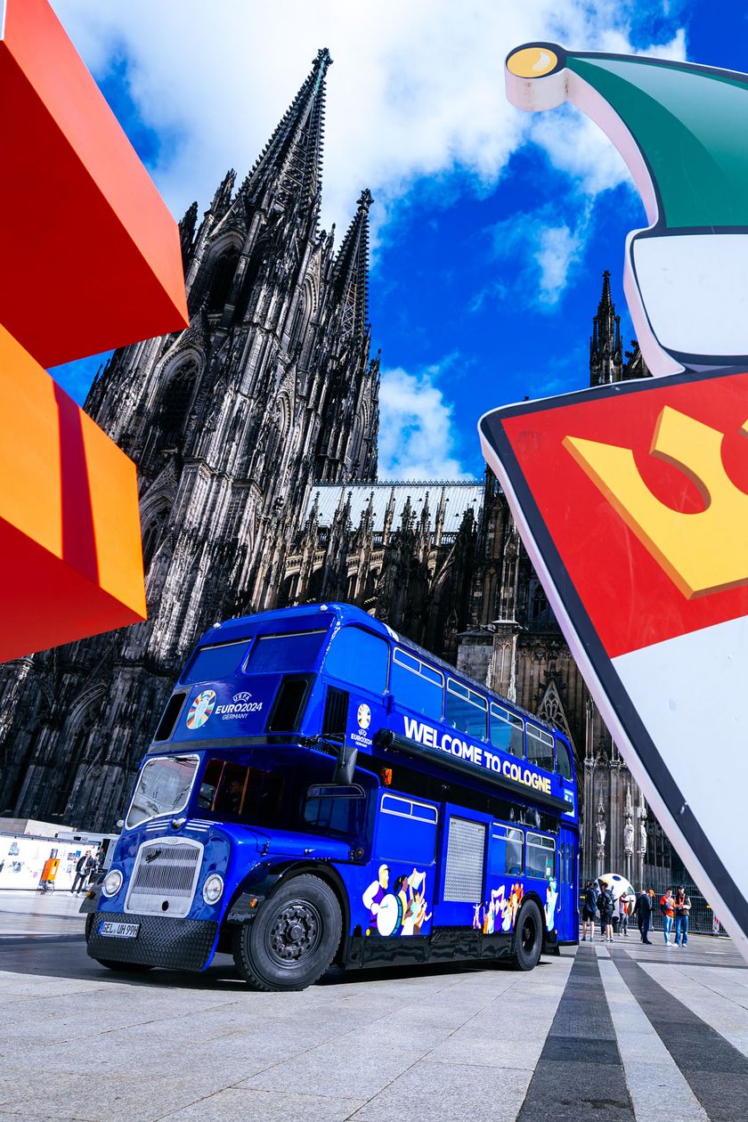 Ein blauer Doppeldeckerbus steht auf dem Platz vor dem Kölner Dom. Zwei Schilder im Vordergrund flankieren das Bild.