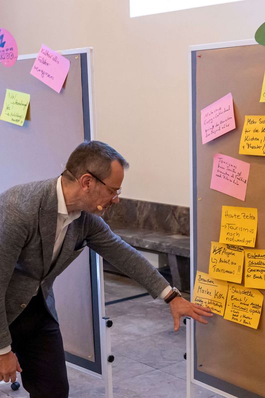 Drei Flipcharts mit beschriebenen Notizzetteln stehen neben einander. Ein Mann ließt eine Zettel vor.