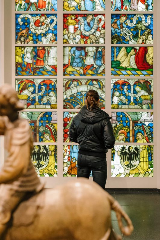 In einem hellen Raum sind christliche Skulpturen arrangiert. Eine Person schaut sich ein großes Buntglasfenster an. 