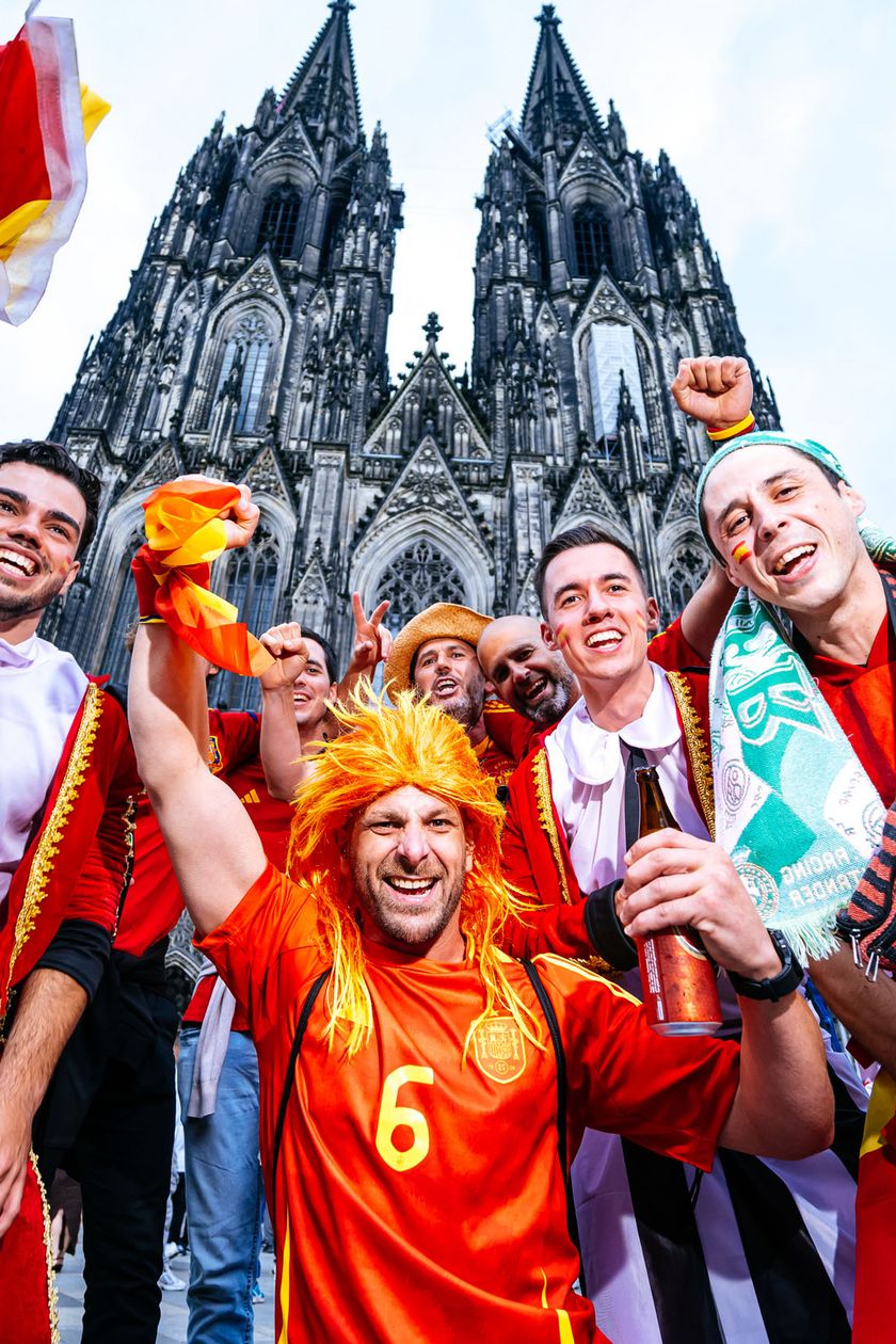 Eine Gruppe von Männern teils in roten Trikots oder verkleidet mit Perücke posieren jubelnd vor dem Kölner Dom.