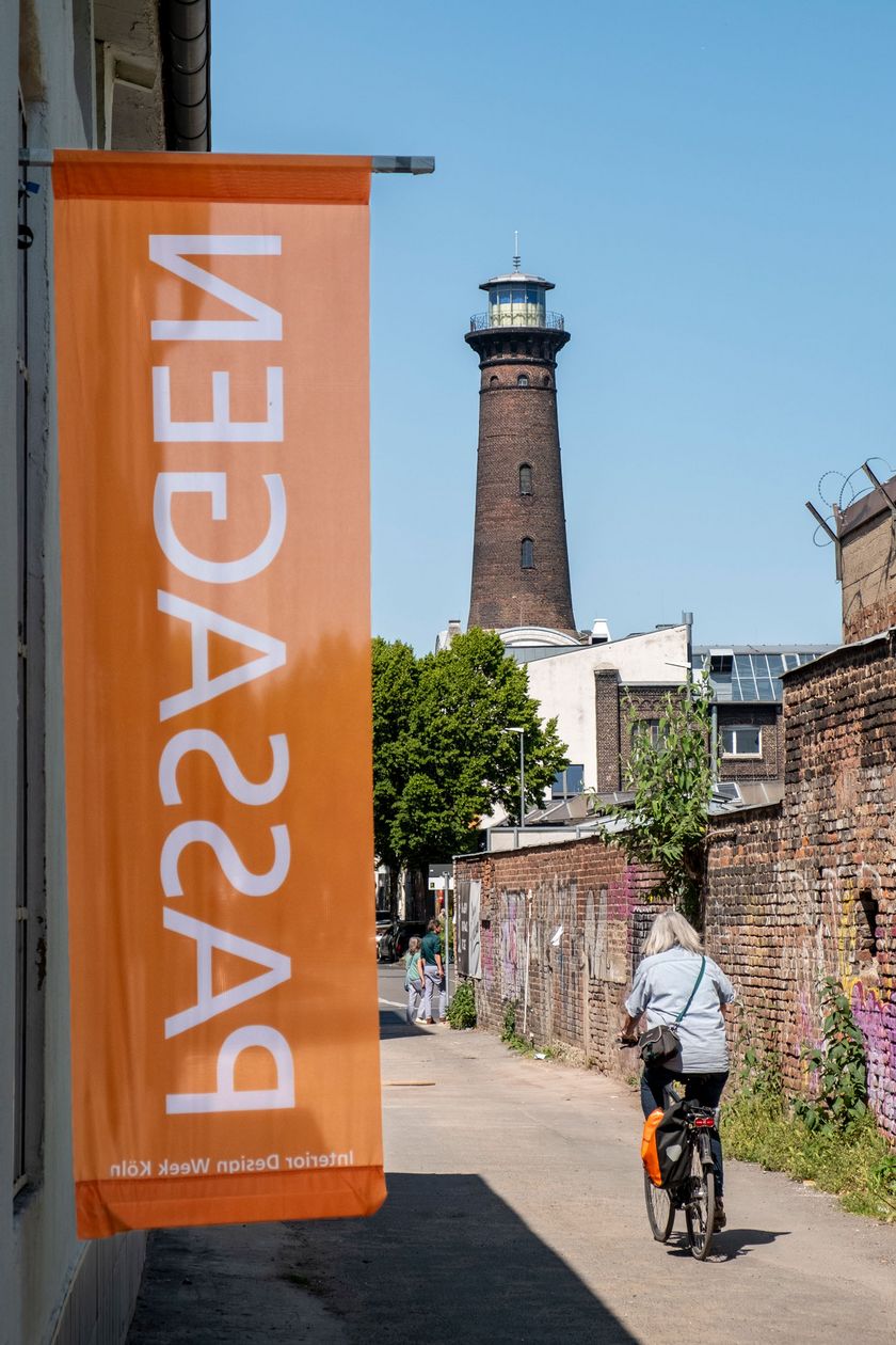 Auf dem Bild ist eine Seitenstraße mit Backsteinmauer zu sehen. Eine Fahne mit der Aufschrift PASSAGEN sticht präsent hervor. Ein Leuchtturm ragt im Hintergrund empor.