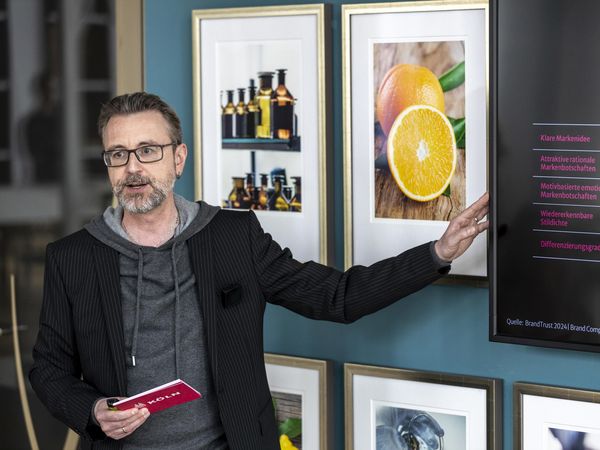 Herr Dr. Amann hält einen Vortrag neben einem digitalen Bildschirm. Die Wände sind mit farbenfrohen gerahmten Bildern dedkoriert.