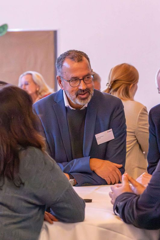 Eine Gruppe von Menschen stehen zusammen um einen runden Stehtisch und führen eine anregend fröhliche Unterhaltung. 